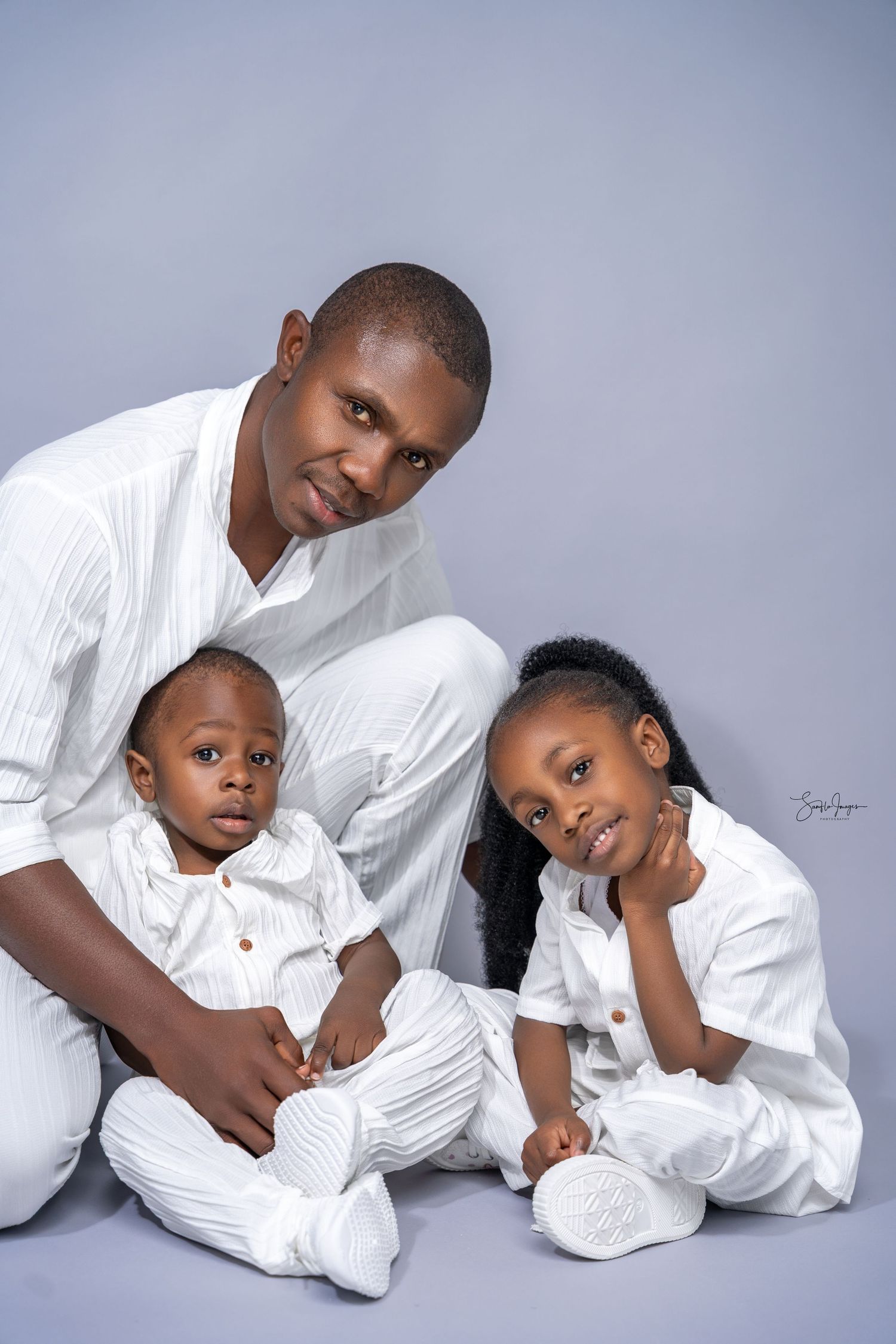 A man and two young children in matching white outfits pose together for a studio family portrait.