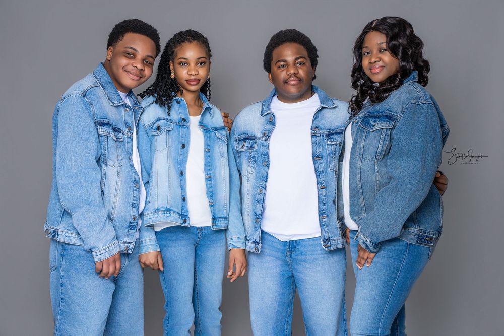 Four people in matching denim jackets and jeans pose together for a coordinated family portrait against a gray background.