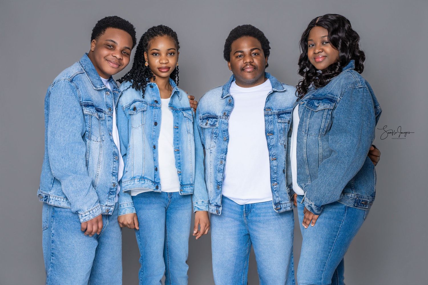 Four people in matching denim jackets and jeans pose together for a coordinated family portrait against a gray background.