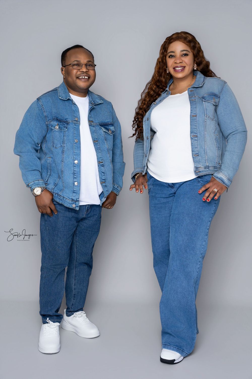 Two people in matching denim jackets and jeans pose together smiling against a gray studio background.