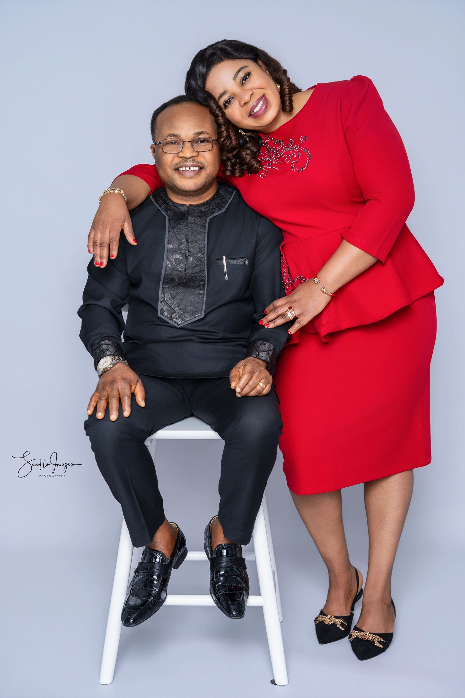 A man in black traditional attire sits while a woman in a red dress leans on his shoulder in a studio portrait.