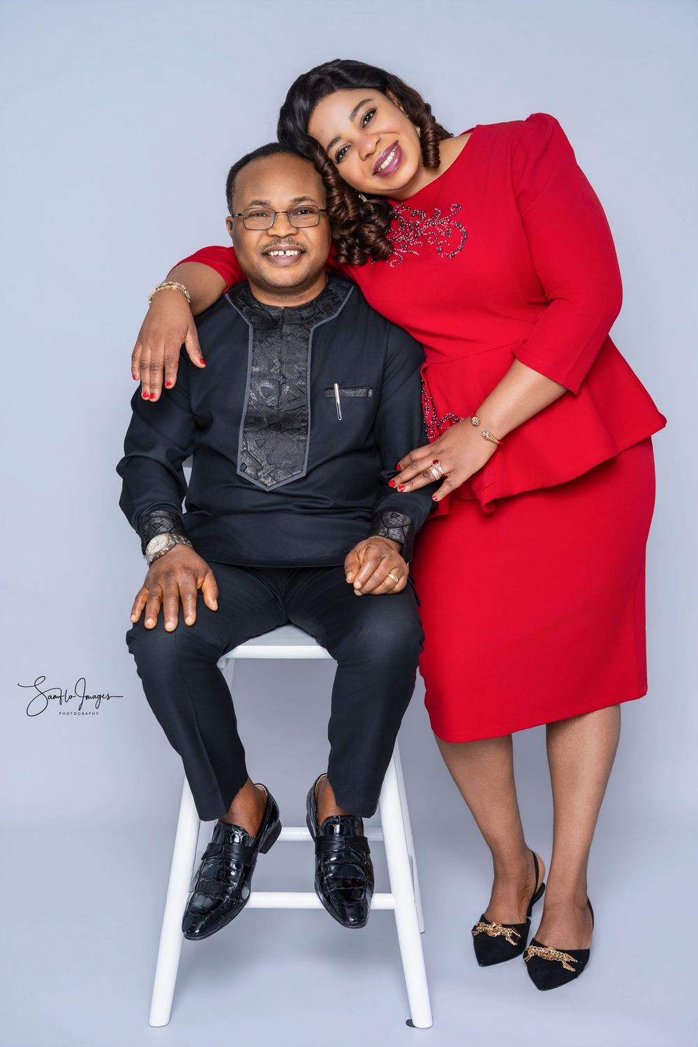 A man in black traditional attire sits while a woman in a red dress leans on his shoulder in a studio portrait.