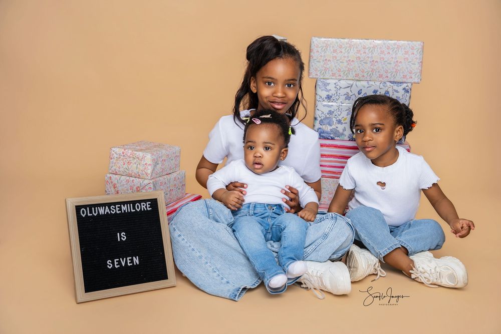 Three children pose for a seventh birthday photo with gift boxes and a letter board reading Oluwasemilore Is Seven.