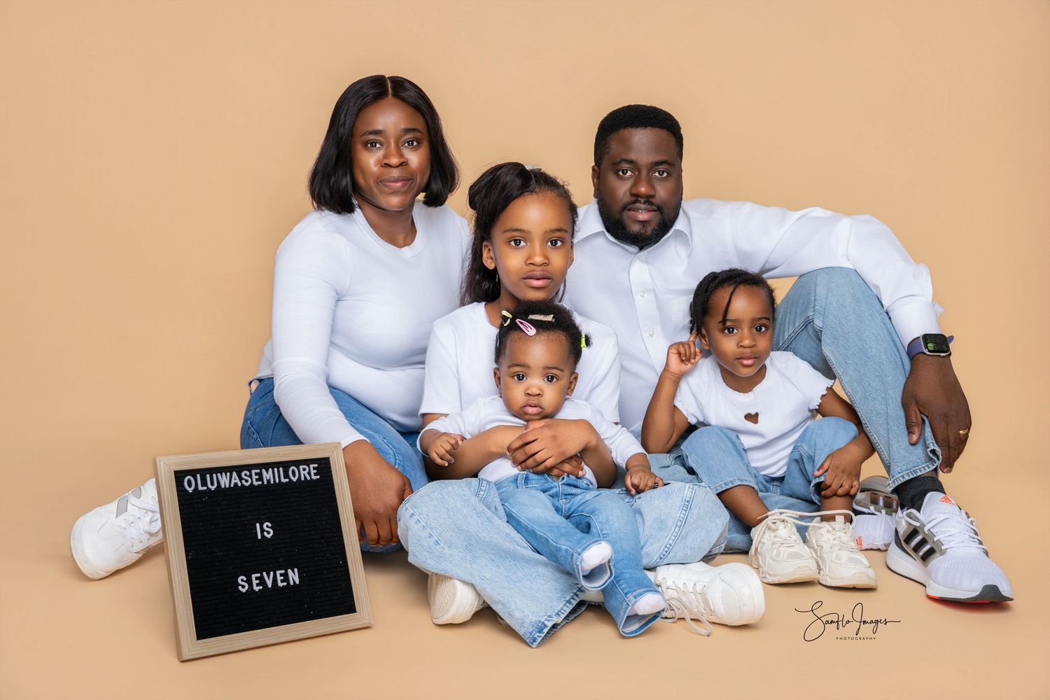 Family of five posing together for a professional portrait with a letter board reading Oluwasenilore is Seven.