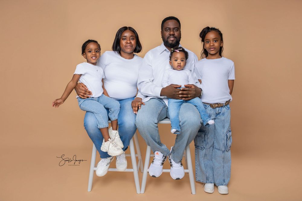 Family of five in matching white tops and jeans poses for a studio portrait against a warm beige background.