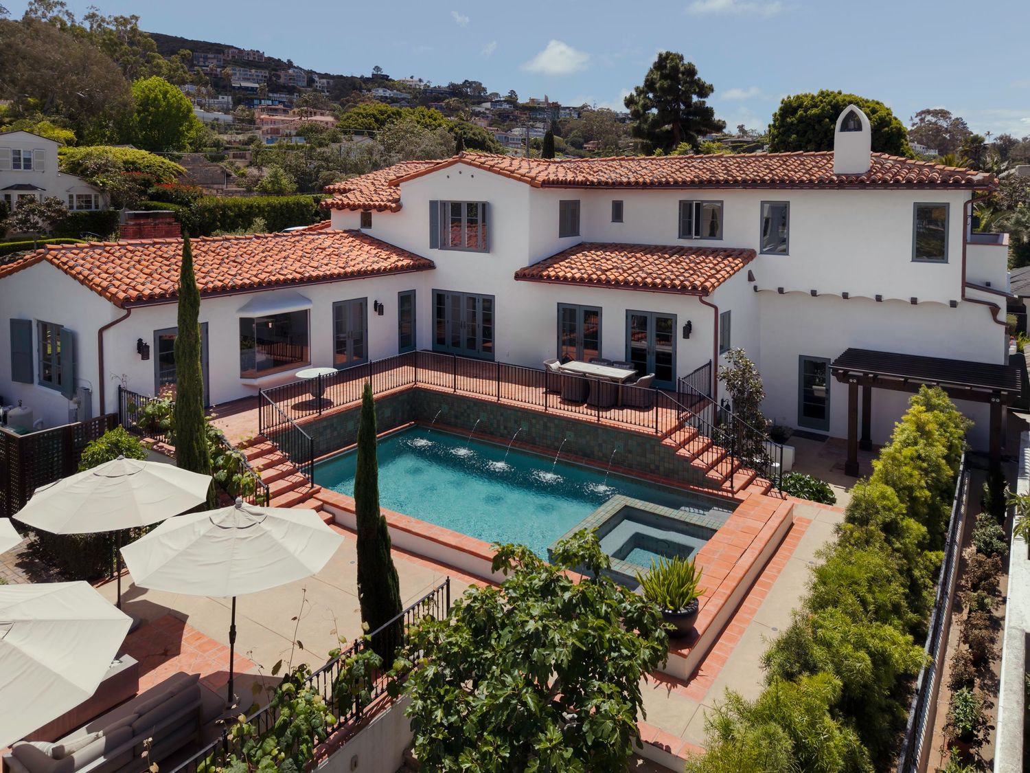 Aerial view of a Spanish-style white stucco home with red tile roof, outdoor pool, spa, and patio in a hillside neighborhood.