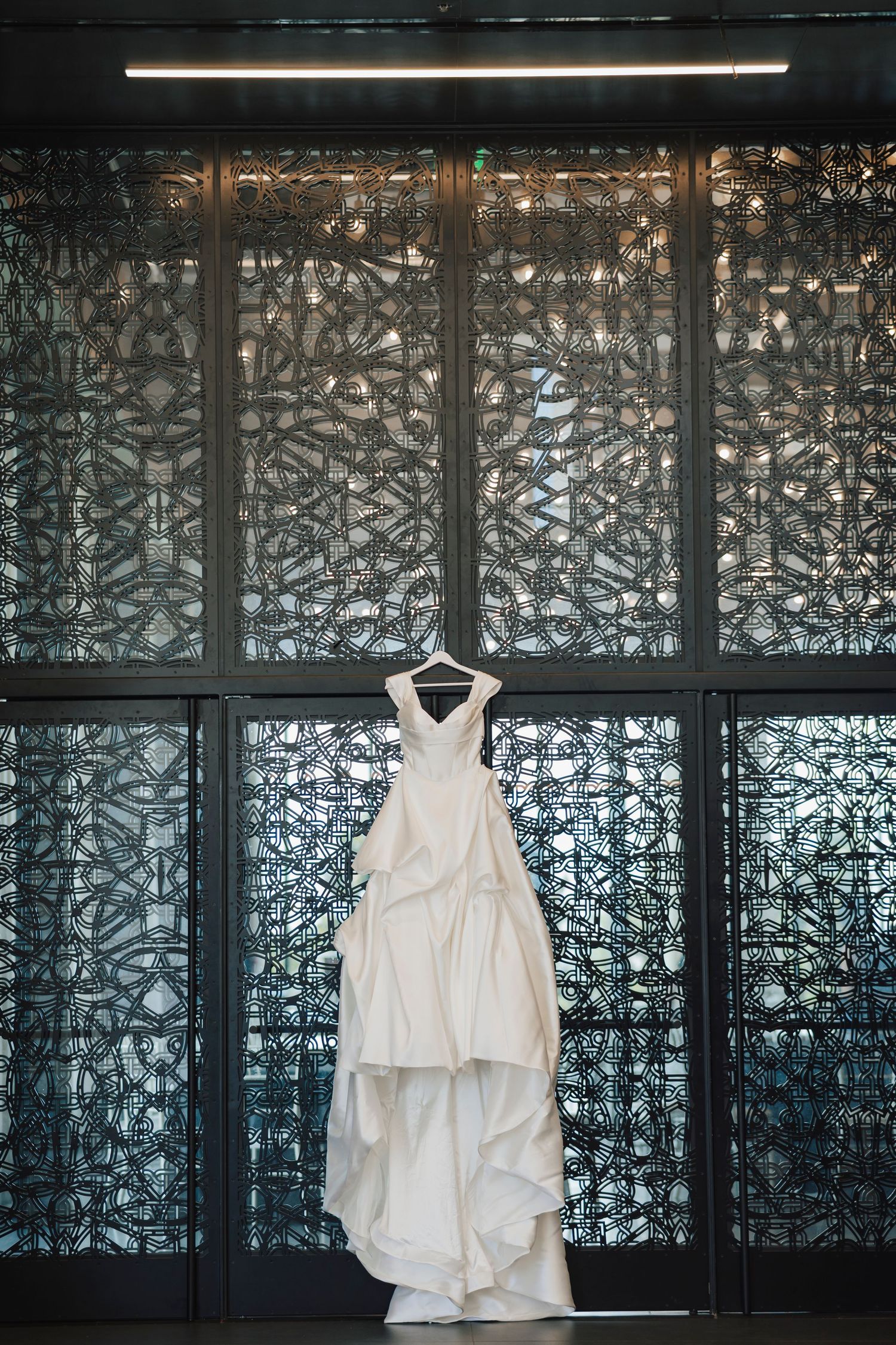 Elegant white wedding gown displayed on a hanger against an ornate decorative metal panel backdrop.