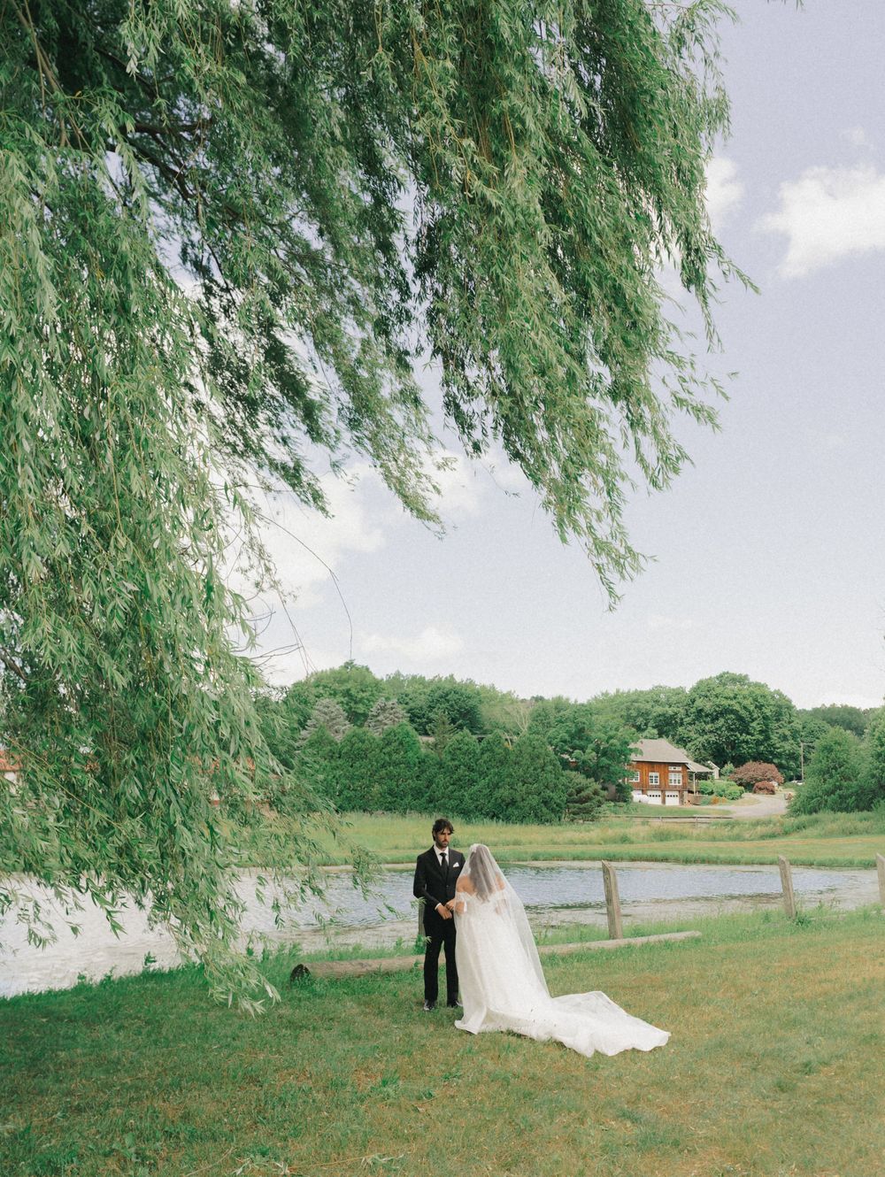 Bride and groom stand beneath a weeping willow tree by a pond in a lush green countryside wedding setting.