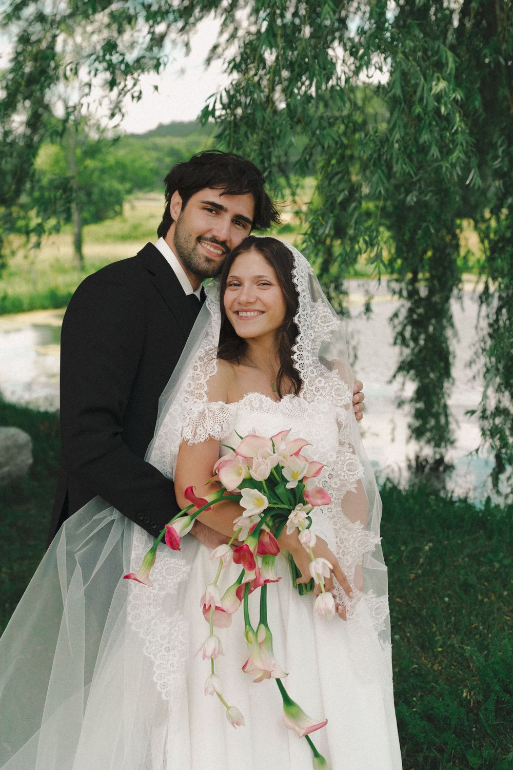 Smiling bride and groom pose under willow tree, bride holding pink tulip bouquet in white lace gown with veil.