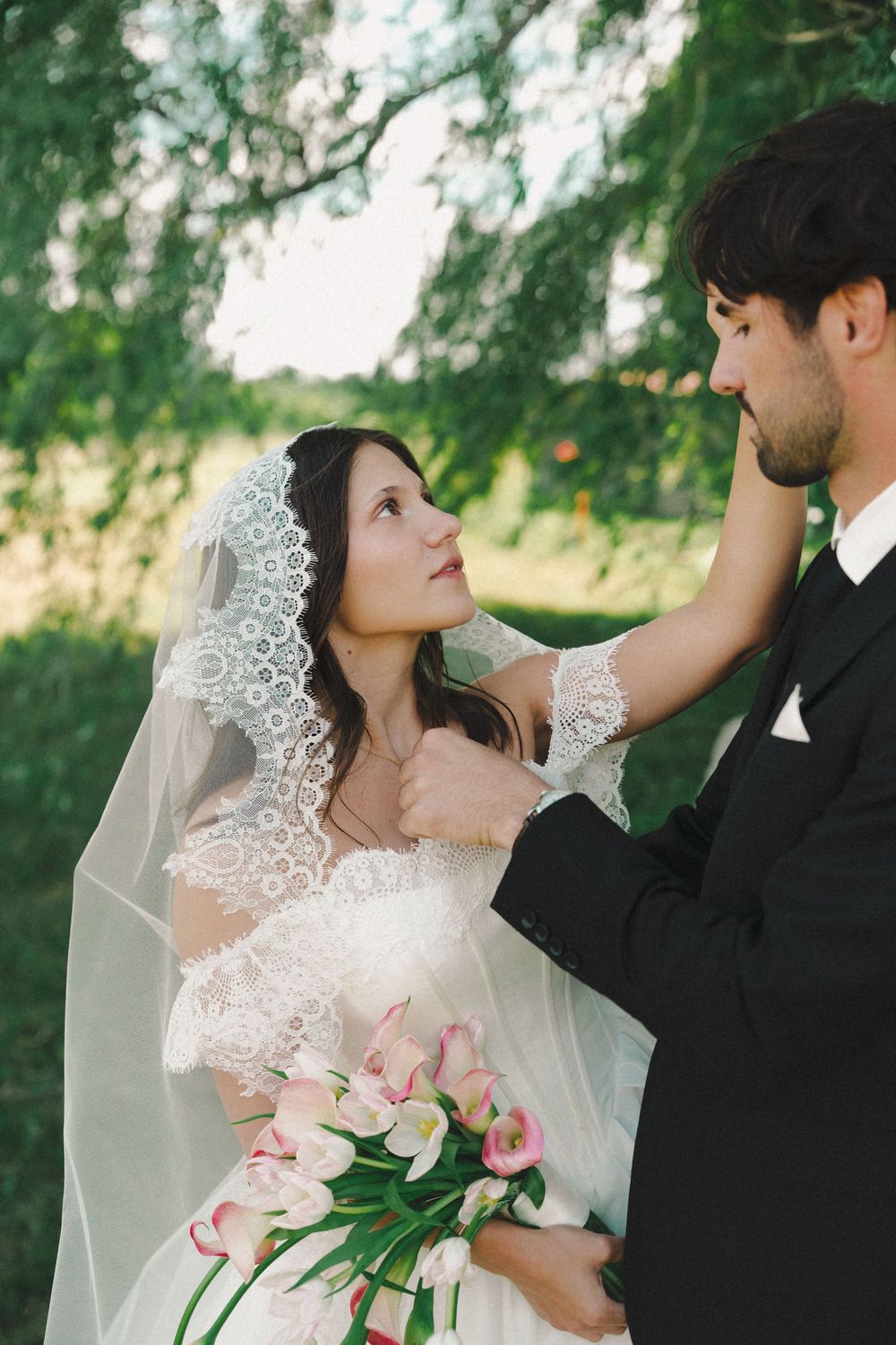 Bride in lace veil holding pink tulips gazes at groom in black suit outdoors.