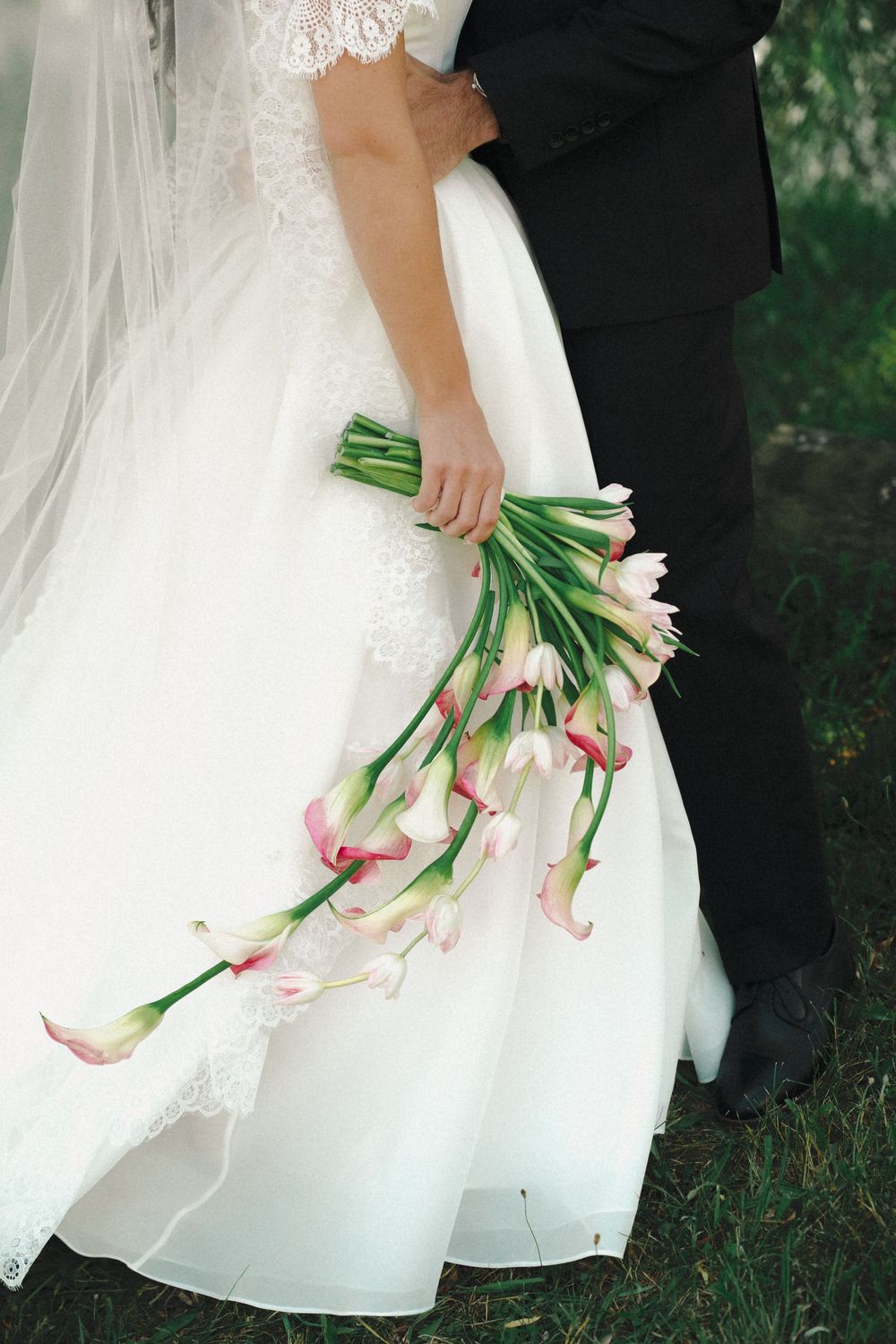 Bride in white lace gown holding elegant pink calla lily bouquet while standing with groom in black suit outdoors.