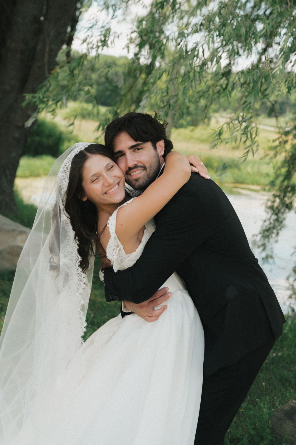 Happy bride and groom embrace outdoors near willow tree, bride in lace veil and white gown, groom in black suit.