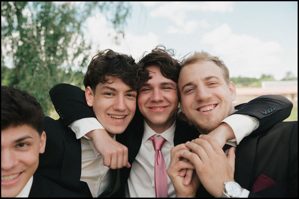 Four young men in suits smiling and posing together outdoors at a formal event.