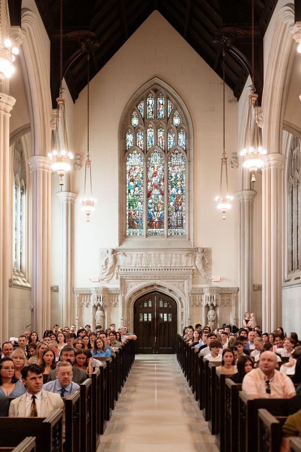 Guests fill wooden pews inside a grand Gothic church with soaring arched ceilings and colorful stained glass windows.