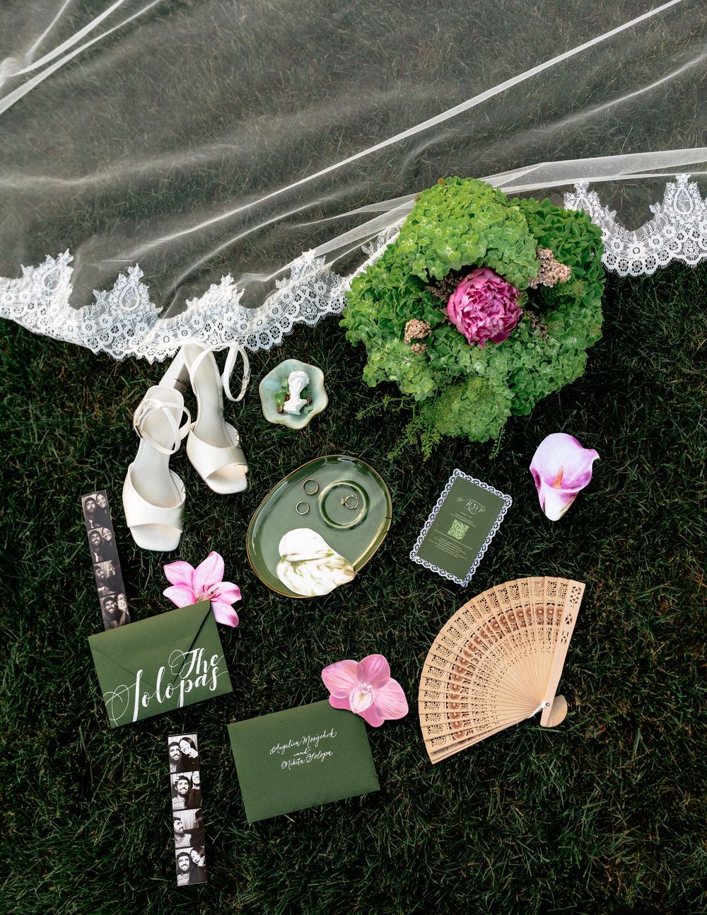 Elegant wedding flat lay with white heels, green stationery, floral bouquet, veil, and decorative accessories on grass.