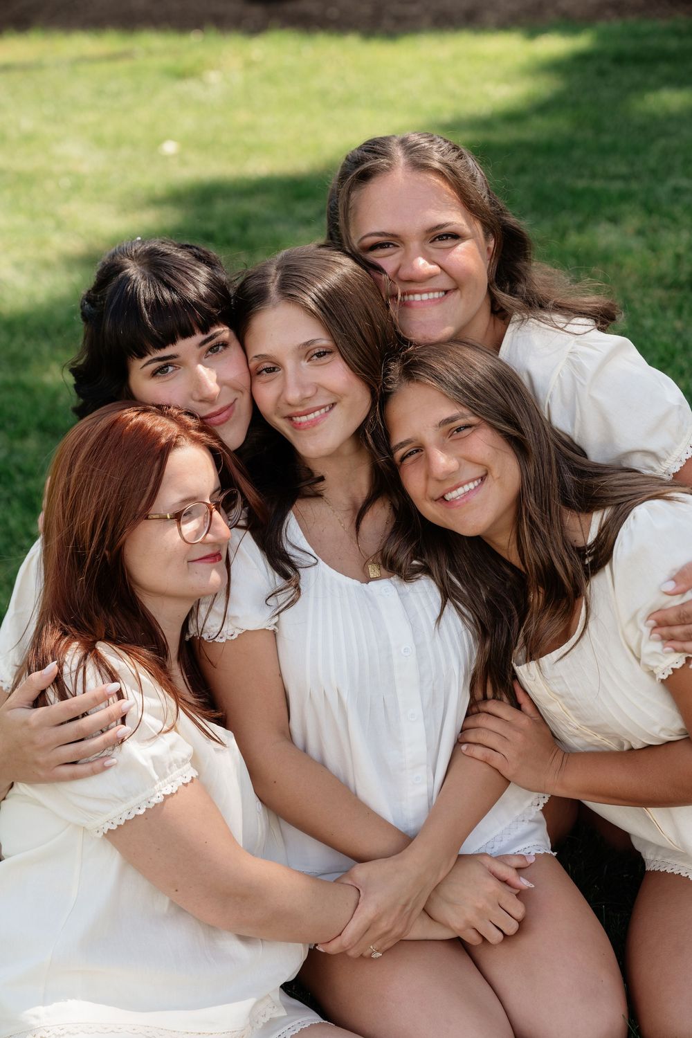 Five young women in white dresses smile and embrace outdoors on green grass in a joyful group portrait.