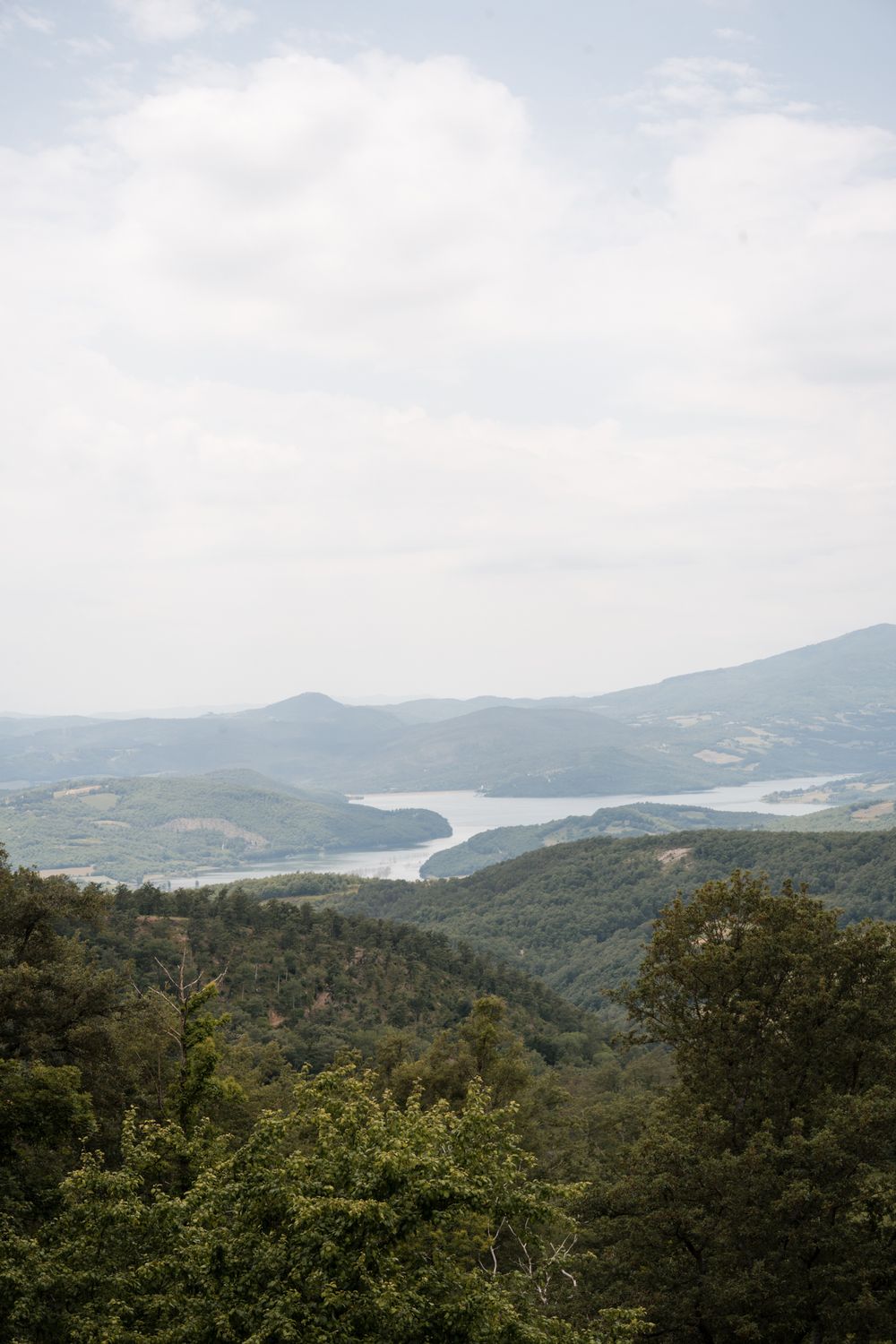 Scenic aerial view of rolling green Tuscan hills with a serene lake and cloudy sky in the background.