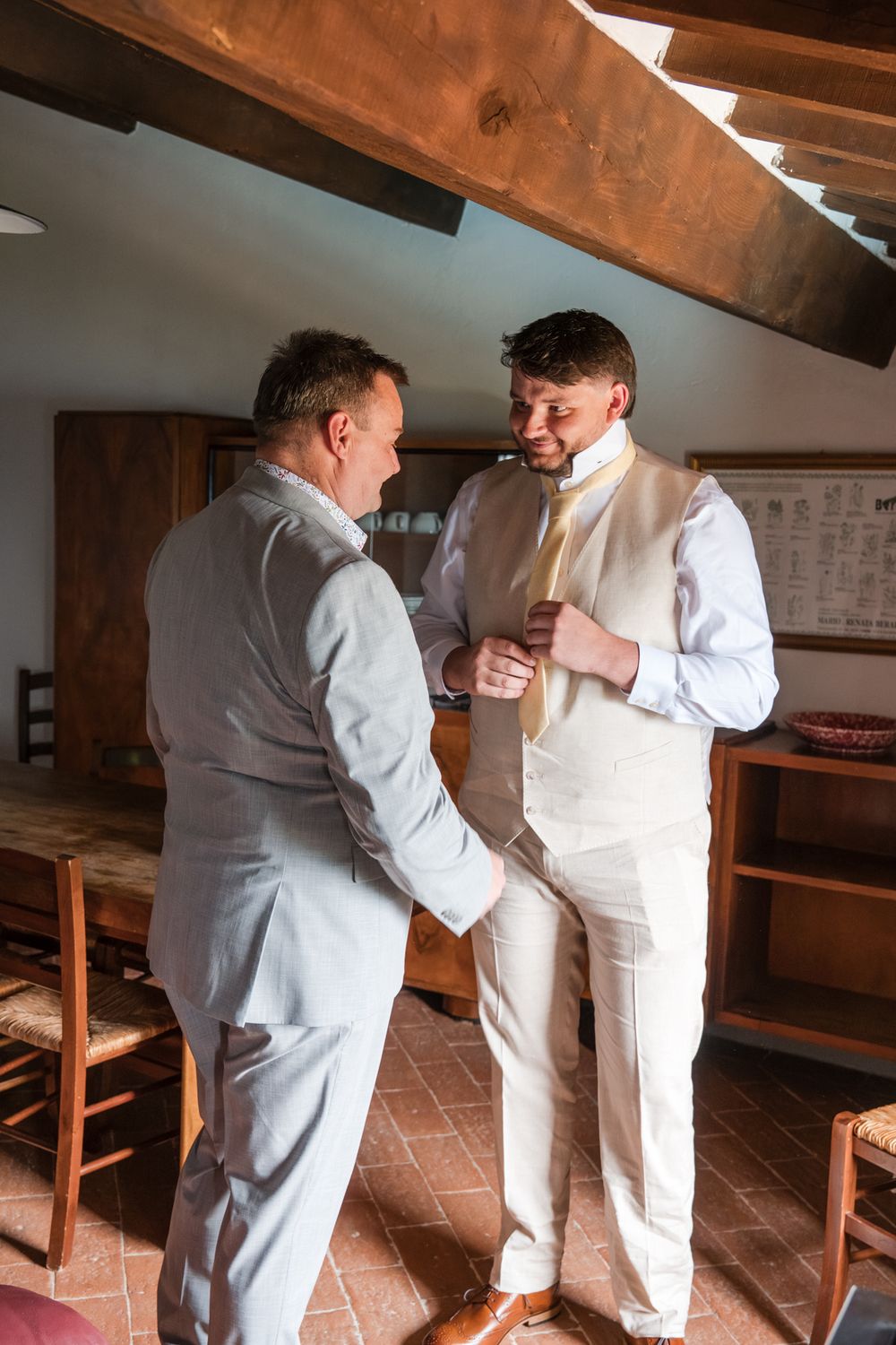 Two men in light suits sharing a moment as one helps the other button his vest in a rustic room.