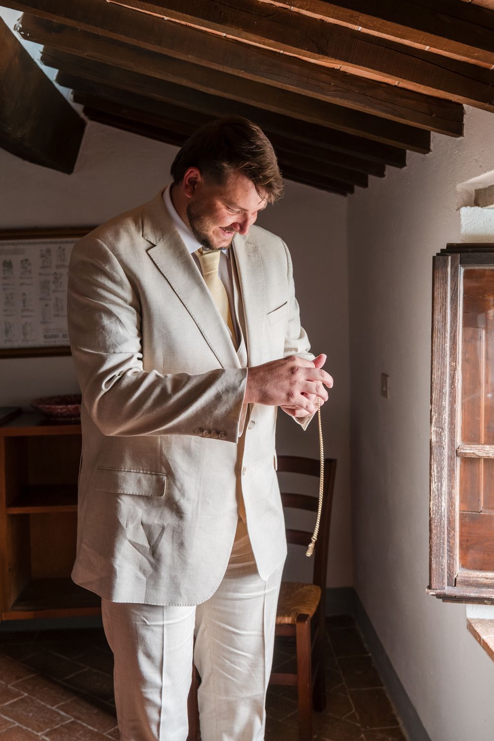 Groom in beige linen suit adjusting his tie near a sunlit window in a rustic Italian villa interior.