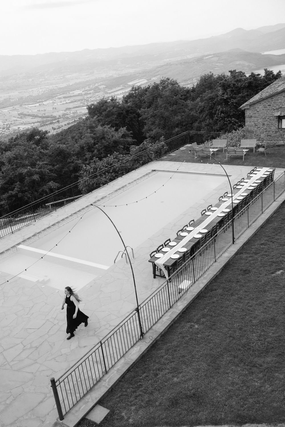 Black and white aerial view of a woman walking past an elegant outdoor wedding reception table setup with scenic valley views.