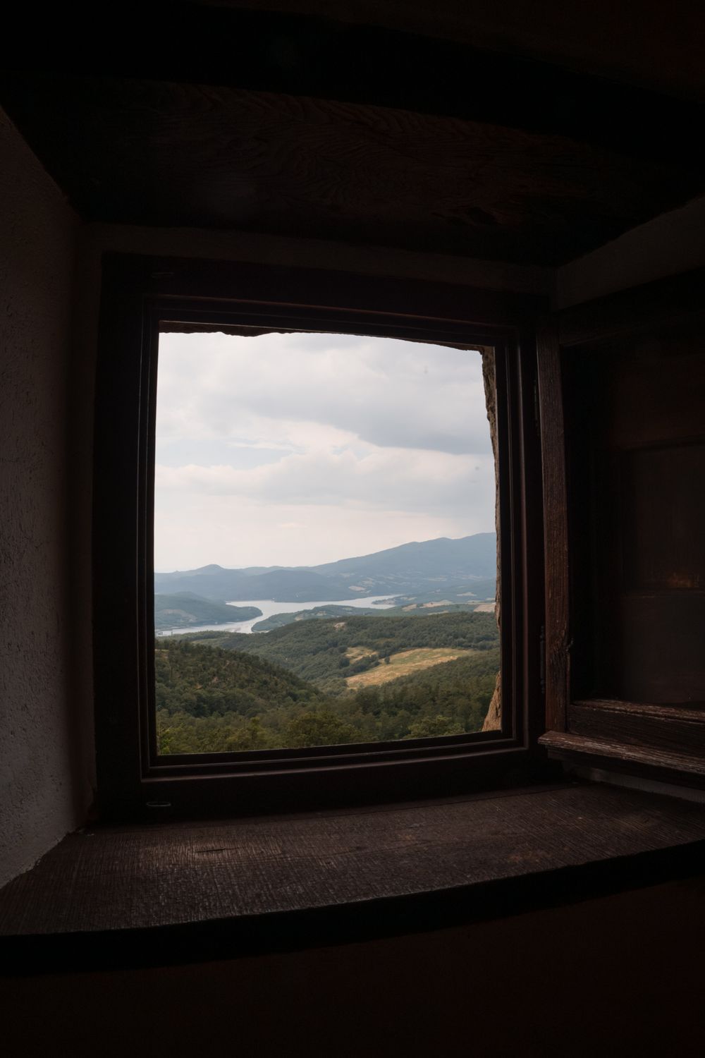 Scenic view through a dark stone window frame of rolling green hills and a lake under a cloudy sky.