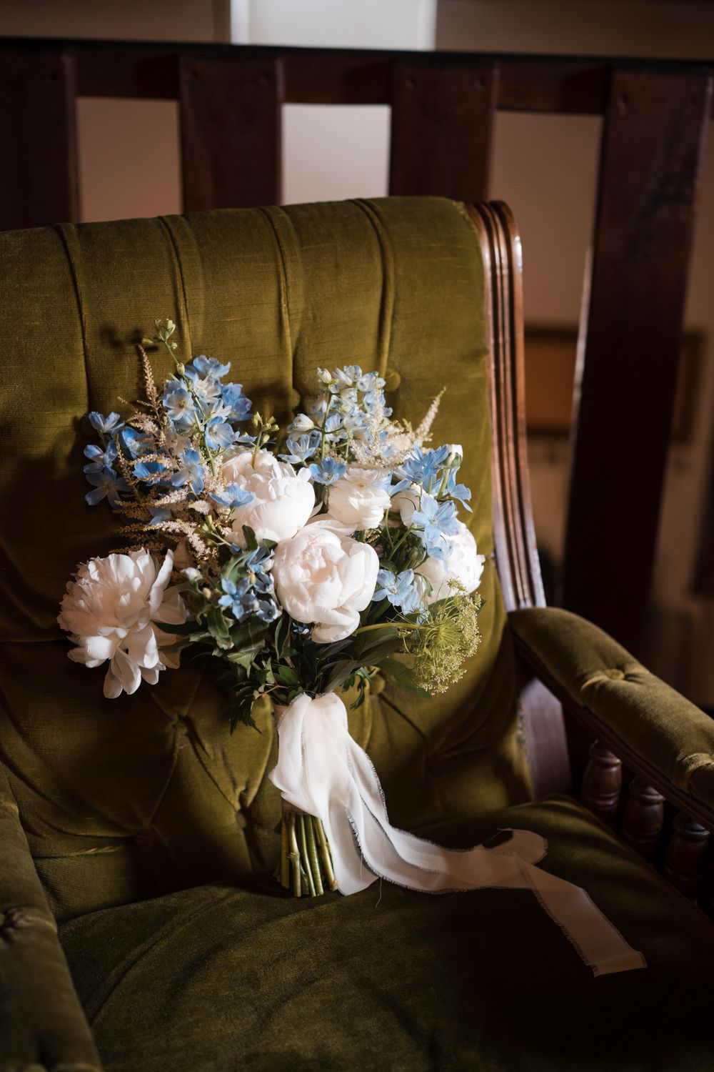Romantic bridal bouquet of white peonies and blue hydrangeas with white ribbon resting on a green velvet armchair.