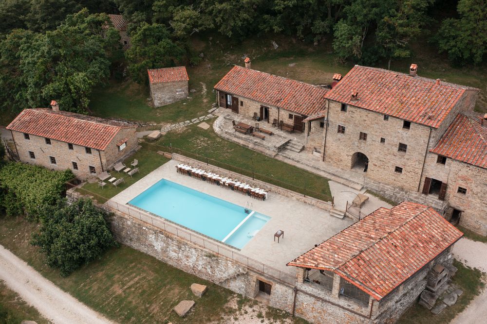Aerial view of a rustic Italian stone farmhouse estate with a rectangular swimming pool surrounded by lush greenery.