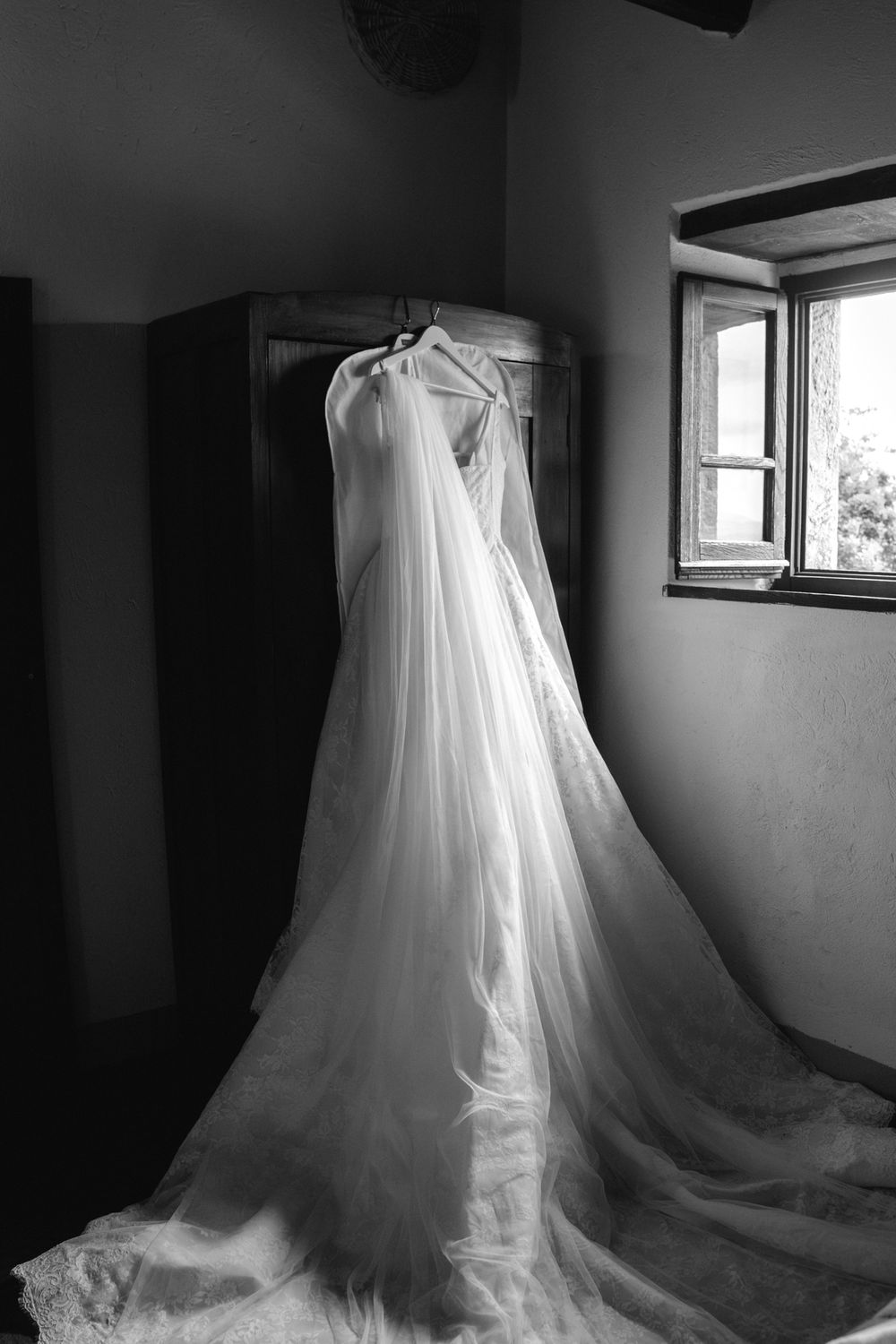 Black and white image of an elegant wedding dress with long veil hanging on a wardrobe near a window.