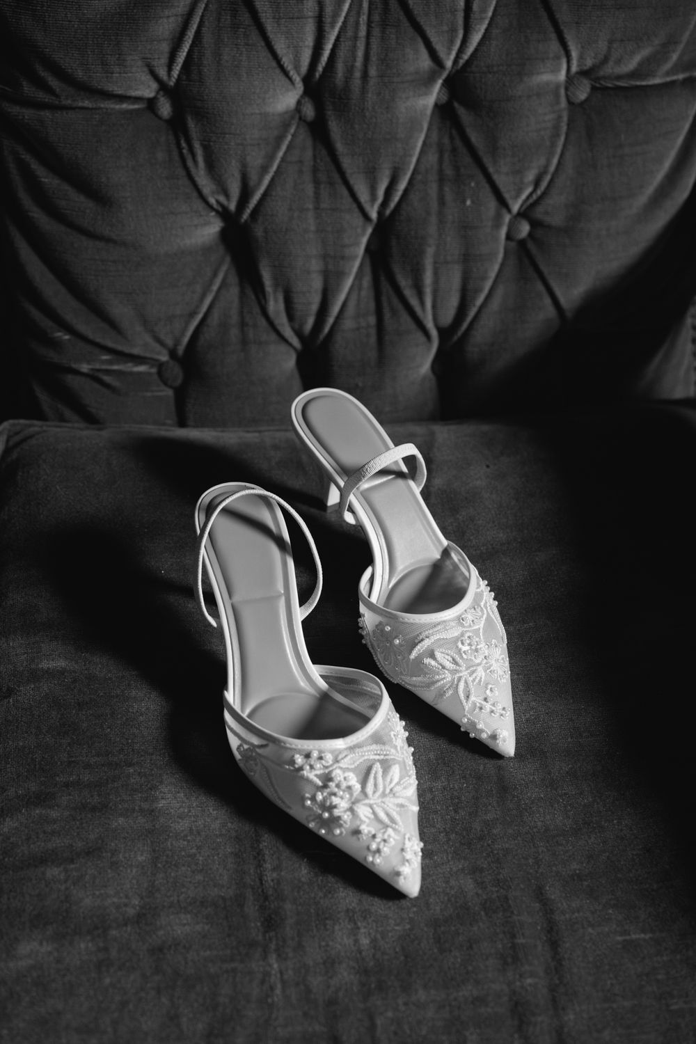Black and white image of embellished bridal heels with floral beading on a tufted velvet chair seat.