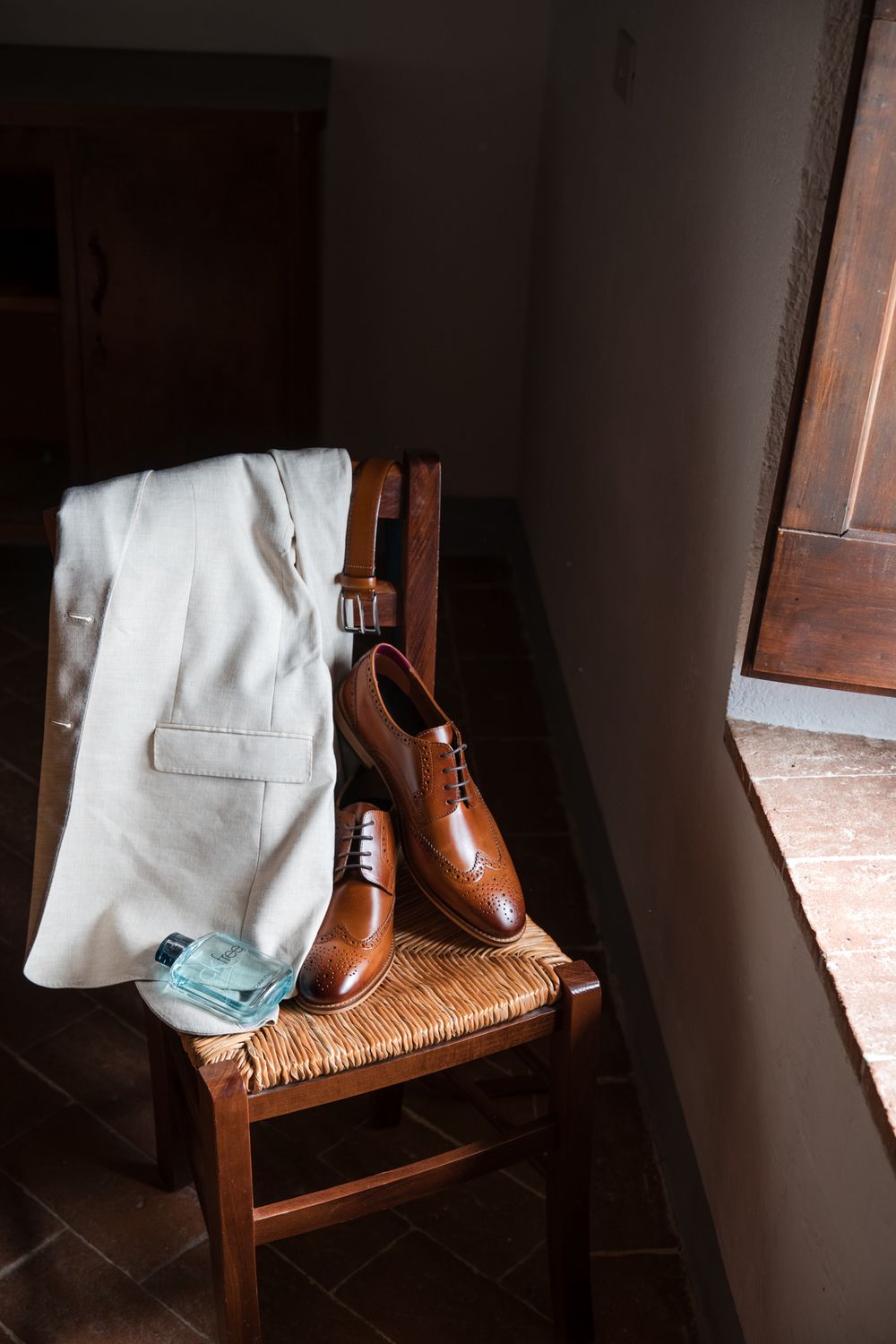 Brown leather Oxford brogue shoes and light suit jacket arranged on a rustic chair with cologne bottle nearby.