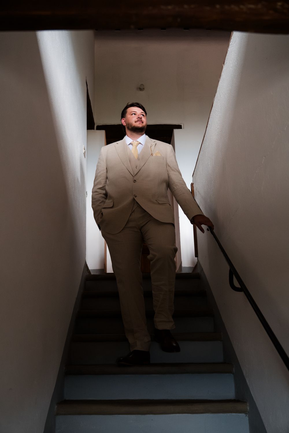 Groom in beige suit descending narrow staircase, low angle black and white portrait.