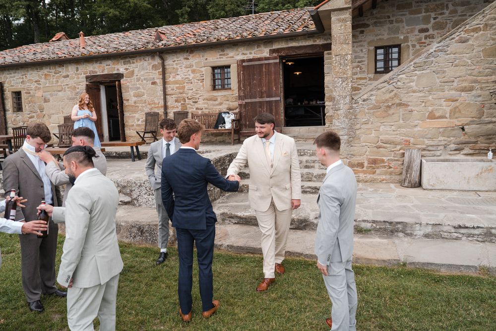 Groom in tan suit greeting groomsmen outside rustic stone venue on wedding day.