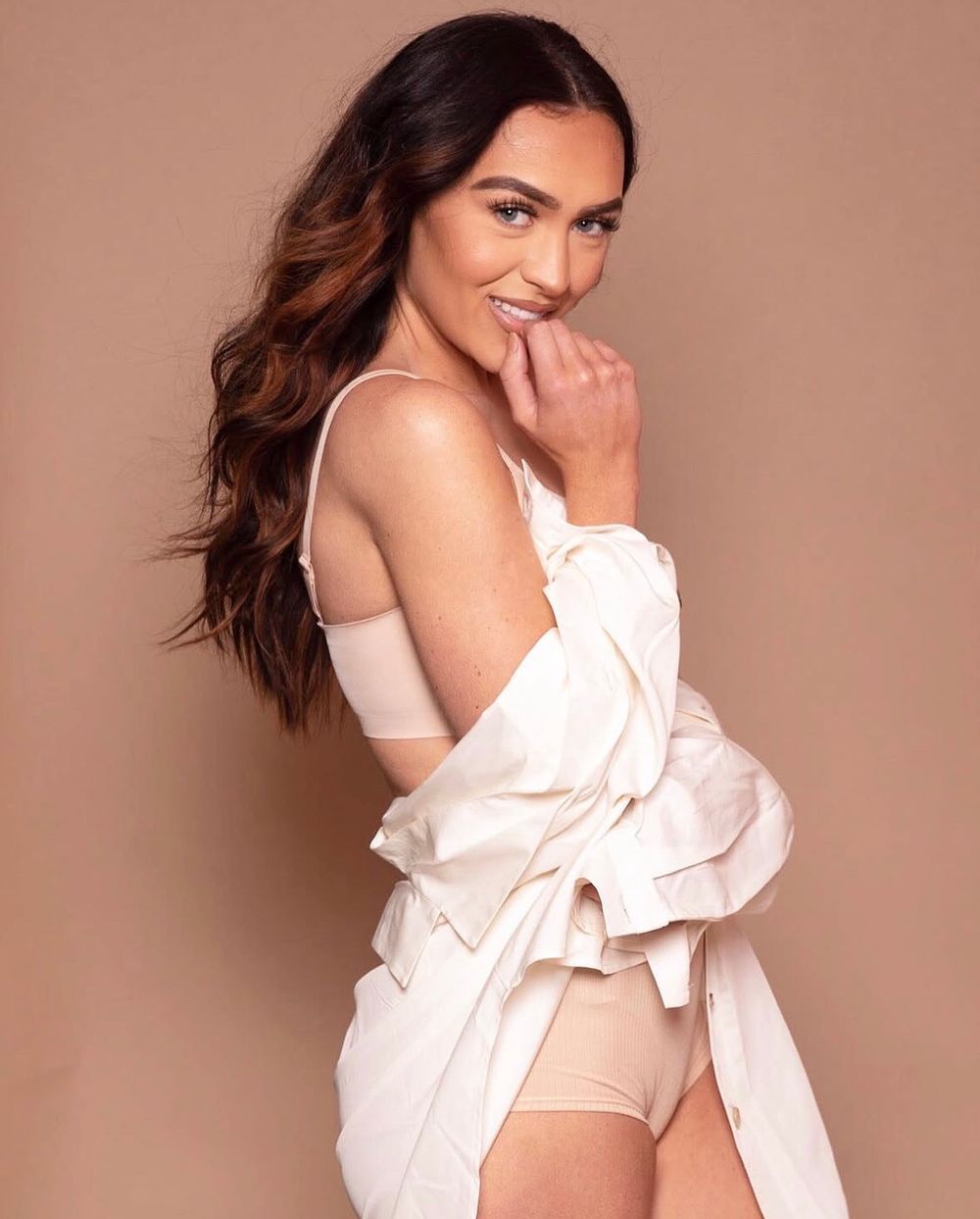 Woman in white shirt and beige underwear smiling over shoulder against tan background.