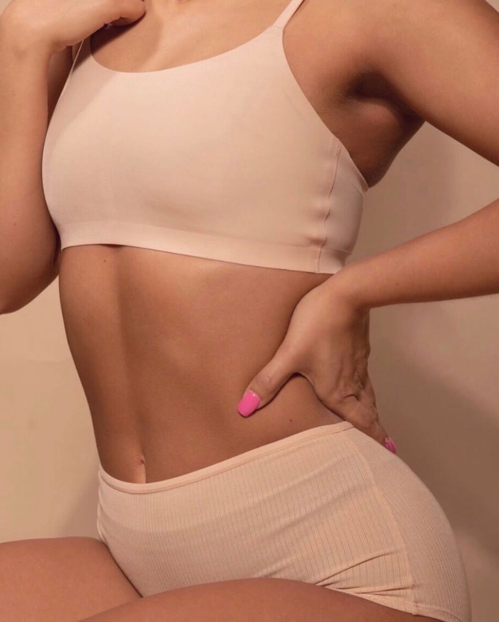 Woman in beige crop top and matching underwear set showcasing a toned midsection with pink manicured nails.