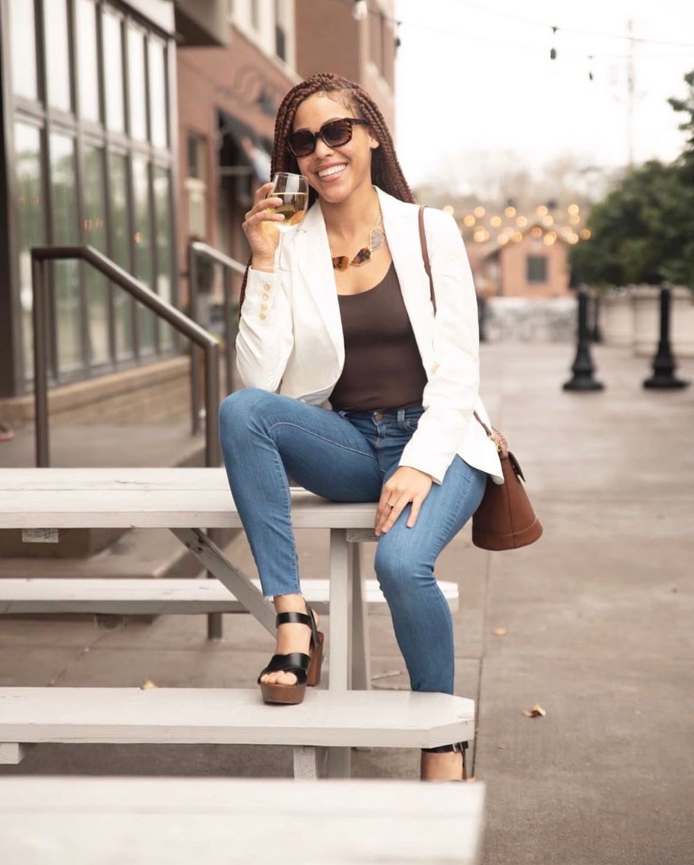 Smiling woman in white blazer, jeans, and sandals sitting on bleachers outdoors, holding a glass of wine.