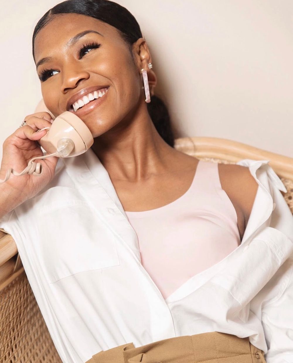Smiling woman in white shirt and pink bodysuit holding a beige mug while sitting in a wicker chair.