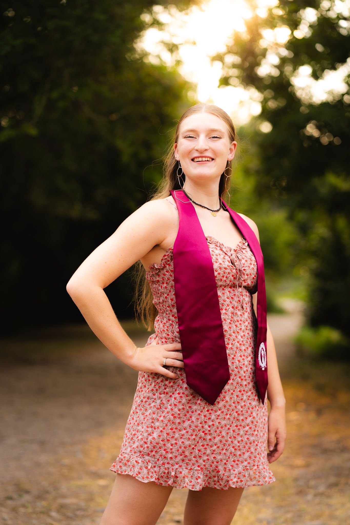 Smiling teenage girl in floral dress and maroon graduation stole poses outdoors in golden hour sunlight.