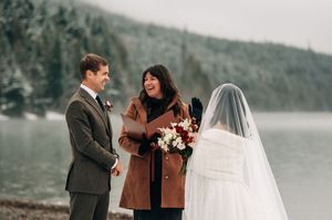 Outdoor winter wedding ceremony by a lake with bride in veil, groom in gray suit, and officiant in brown coat.