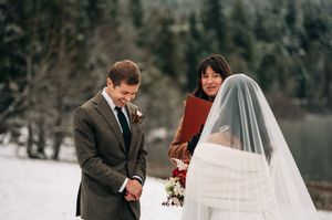 Groom in brown suit reads vows to bride in white dress and veil during snowy winter wedding ceremony outdoors.