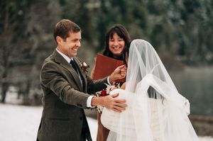 Groom in gray suit smiles at veiled bride during outdoor winter wedding ceremony with officiant holding red book.