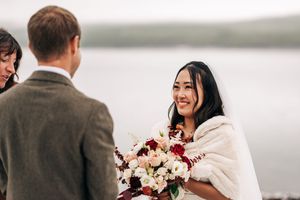Smiling Asian bride holding a bouquet of pink and burgundy flowers during an outdoor waterfront wedding ceremony.