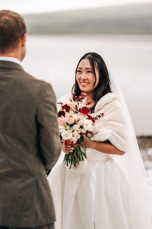 Smiling Asian bride in white gown and fur wrap holds bouquet facing groom outdoors by misty lake.