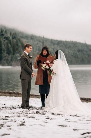 Couple exchanges vows in a snowy winter elopement ceremony by a misty lake with an officiant.