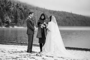 Black and white photo of couple exchanging vows with officiant on a snowy lakeside beach in winter.