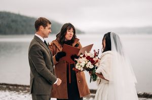 Couple exchanges vows outdoors in winter with officiant by snowy lakeside, bride holding red and white bouquet.