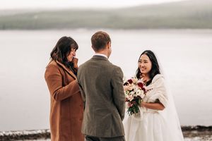Bride smiling at groom during intimate winter elopement ceremony by misty lake with officiant in brown coat.