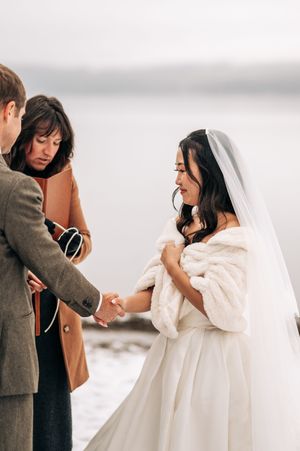 Bride in white fur wrap and veil holds groom's hands during outdoor winter wedding ceremony.