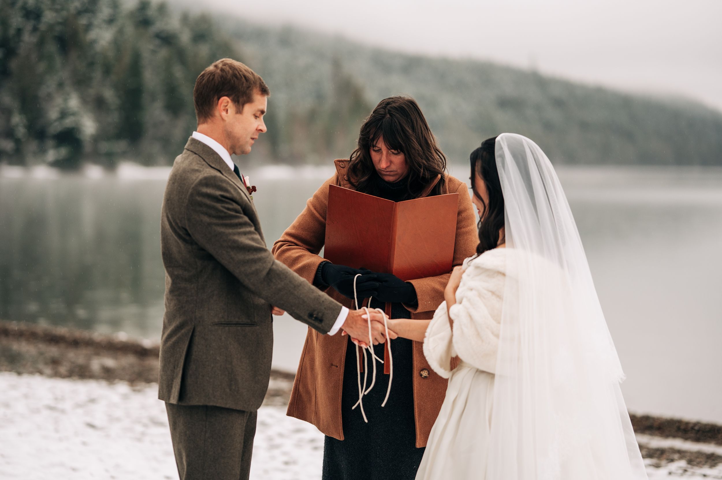 Bride and groom exchange vows outdoors in winter, officiant holding red book by snowy lakeside.