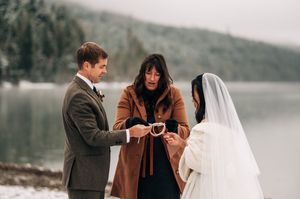 Bride and groom exchange rings with officiant by a snowy lake during an intimate winter wedding ceremony outdoors.