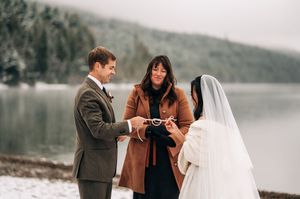 Groom placing ring on brides finger during snowy lakeside elopement ceremony with officiant in brown coat.